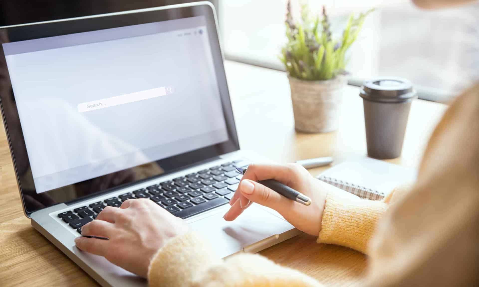 person-typing-on-laptop-displaying-search-engine-bar-at-desk-with-to-go-coffee-cup-and-potted-plant