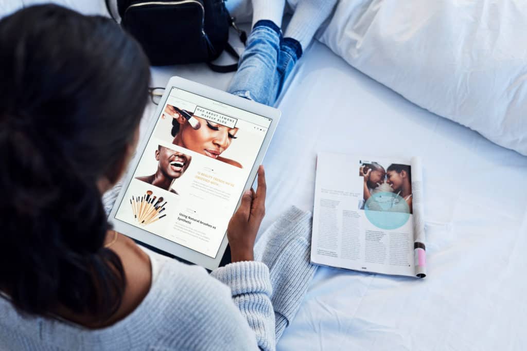 high-angle-shot-of-young-woman-sitting-with-legs-stretched-out-on-bed-while-browsing-future-proof-website-on-tablet-with-magazine-beside-her