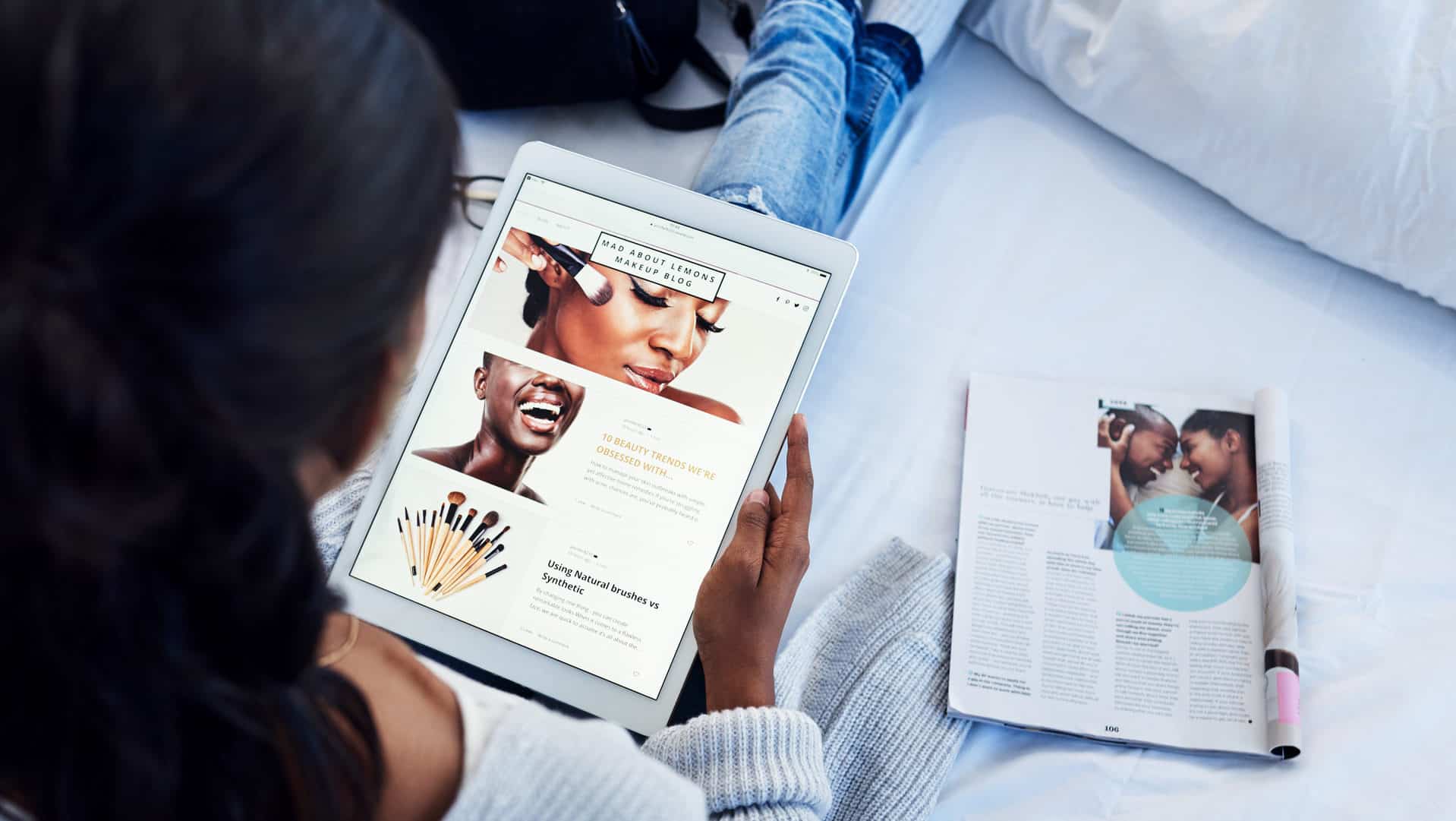 high-angle-shot-of-young-woman-sitting-with-legs-stretched-out-on-bed-while-browsing-website-on-tablet-with-magazine-beside-her